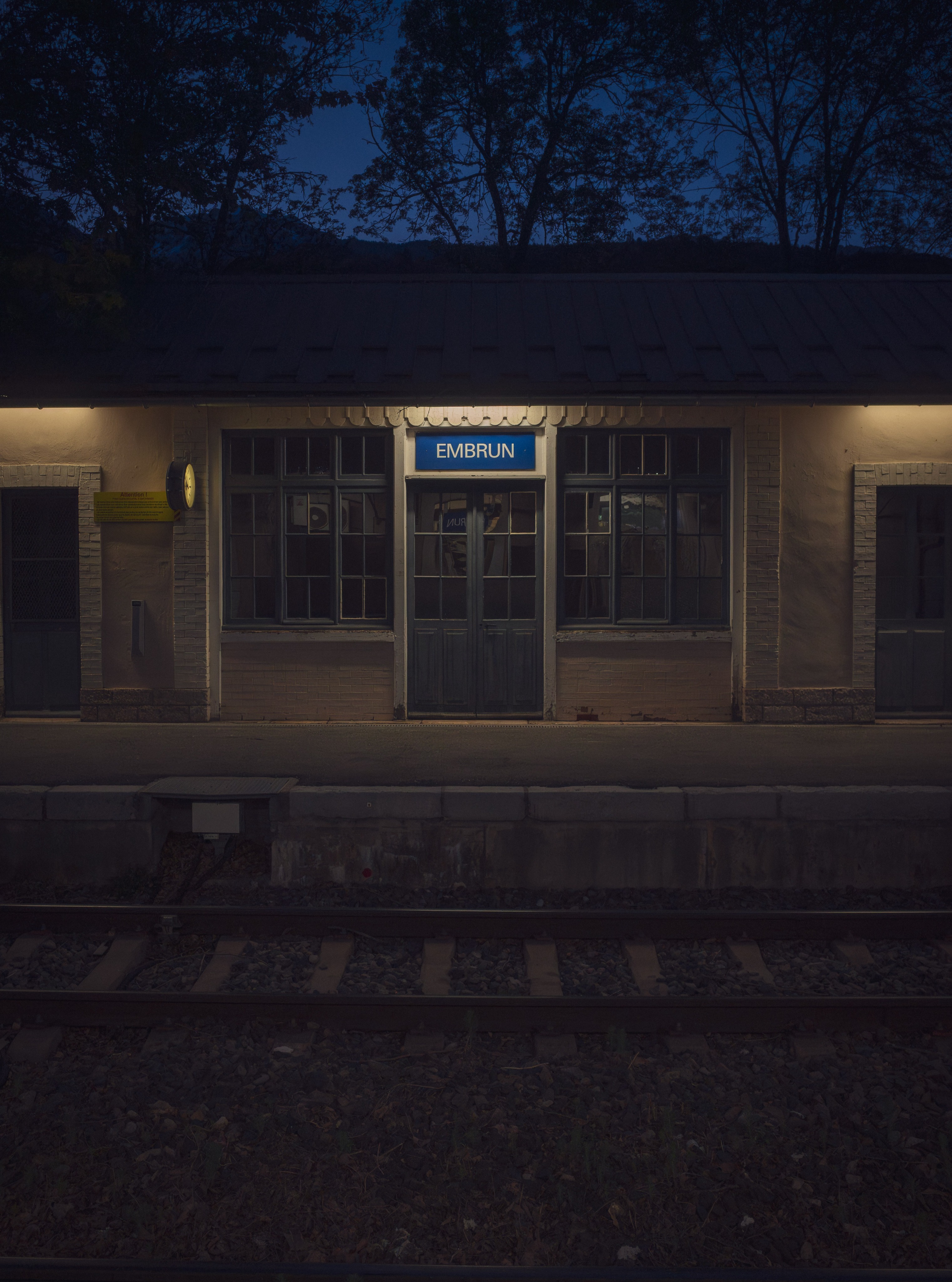 En attendant notre train couchette pour rentrer sur Paris, j’ai pris cette photo sur le quai de la gare d’Embrun, à cette heure que j’aime tant… ce moment suspendu entre le jour et la nuit.

J’ai toujours eu un faible pour les images très symétriques, presque immobiles, et pour ces lumières qui hésitent encore entre le bleu du soir et la chaleur artificielle des quais de gare.

Il y avait quelque chose de calme et de cinématographique dans cette attente. Les rails, le silence, quelques lumières allumées… et cette sensation étrange que certains voyages continuent encore un peu avant même que le train démarre.